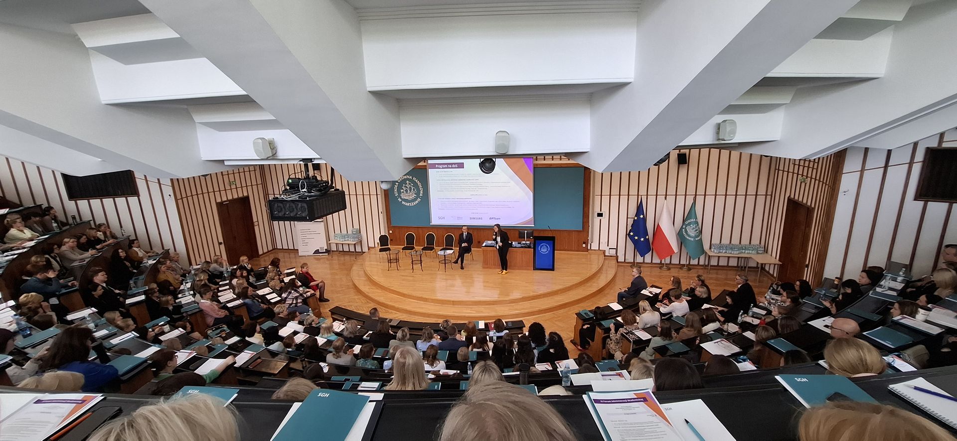 Duża aula wykładowa z widokiem z góry na scenę, gdzie odbywa się prezentacja. Na scenie stoją dwie osoby, a za nimi znajduje się ekran projekcyjny, flagi Unii Europejskiej, Polski i uczelni. Aula jest wypełniona uczestnikami z notatnikami i teczkami.