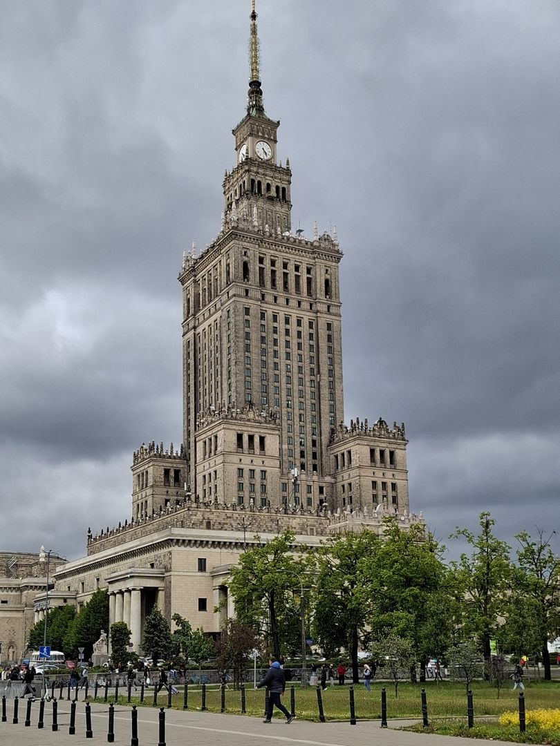 Widok na Pałac Kultury i Nauki w Warszawie w pochmurny dzień. Budynek otoczony jest zielenią i spacerującymi ludźmi. Na pierwszym planie chodnik z czarnymi słupkami.