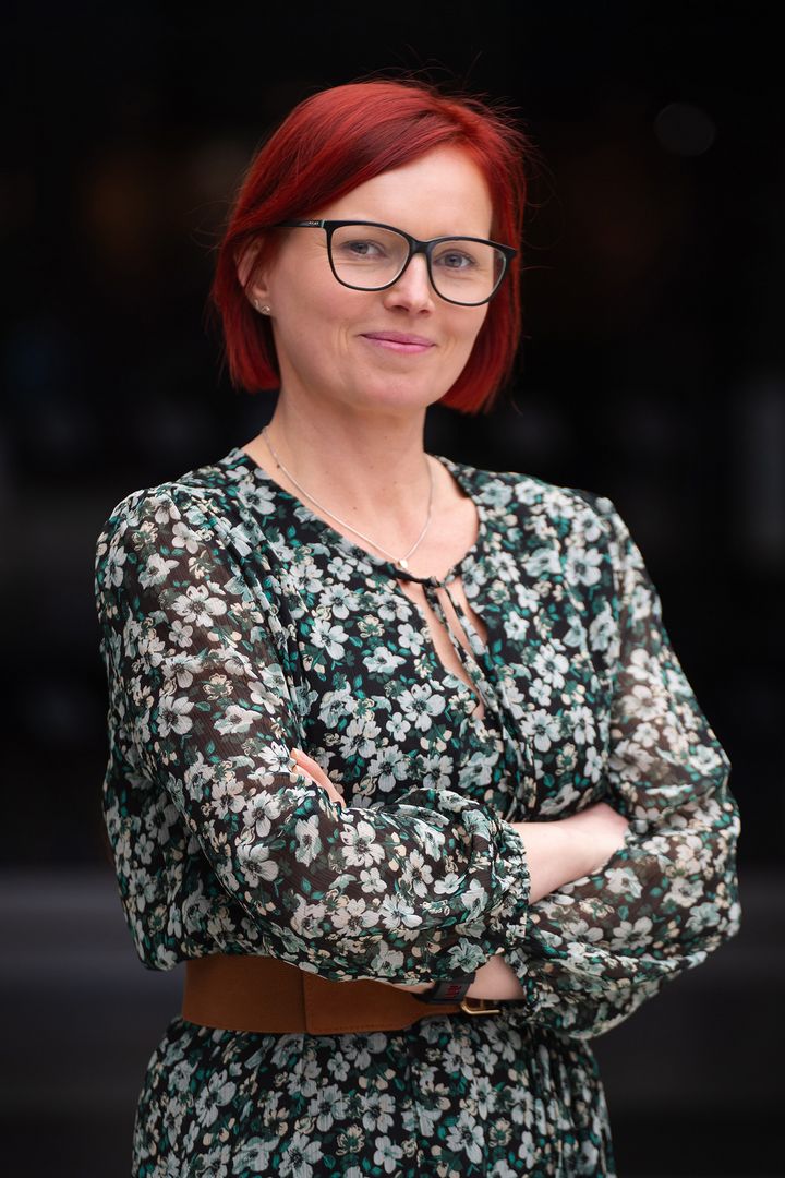 Kobieta z krótkimi, czerwonymi włosami i okularami, uśmiechnięta, w sukience w kwiaty, stoi z założonymi rękami na ciemnym tle.