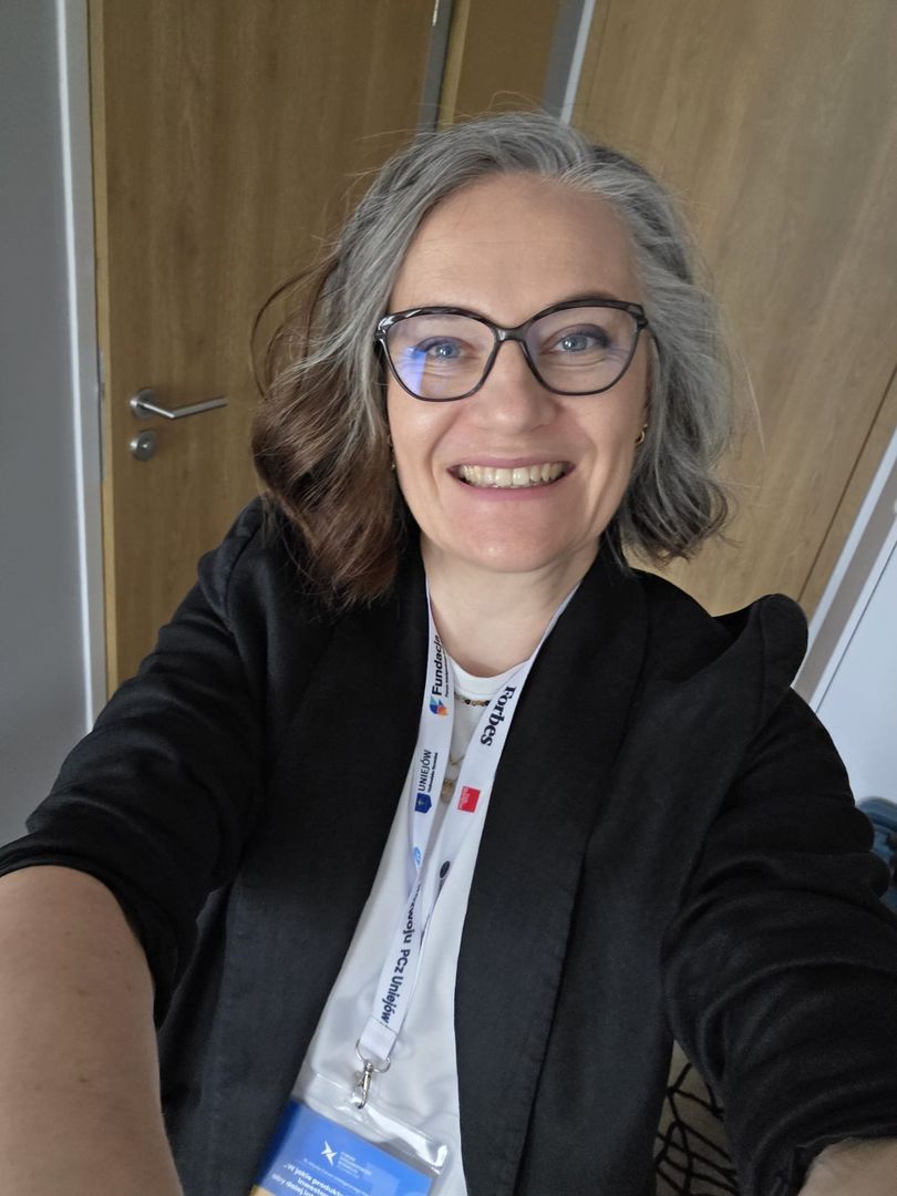 Osoba w czarnej marynarce i białej koszuli robiąca selfie; na szyi zawieszony identyfikator i smycz z logotypami, w tle drewniane drzwi i fragment ściany. 