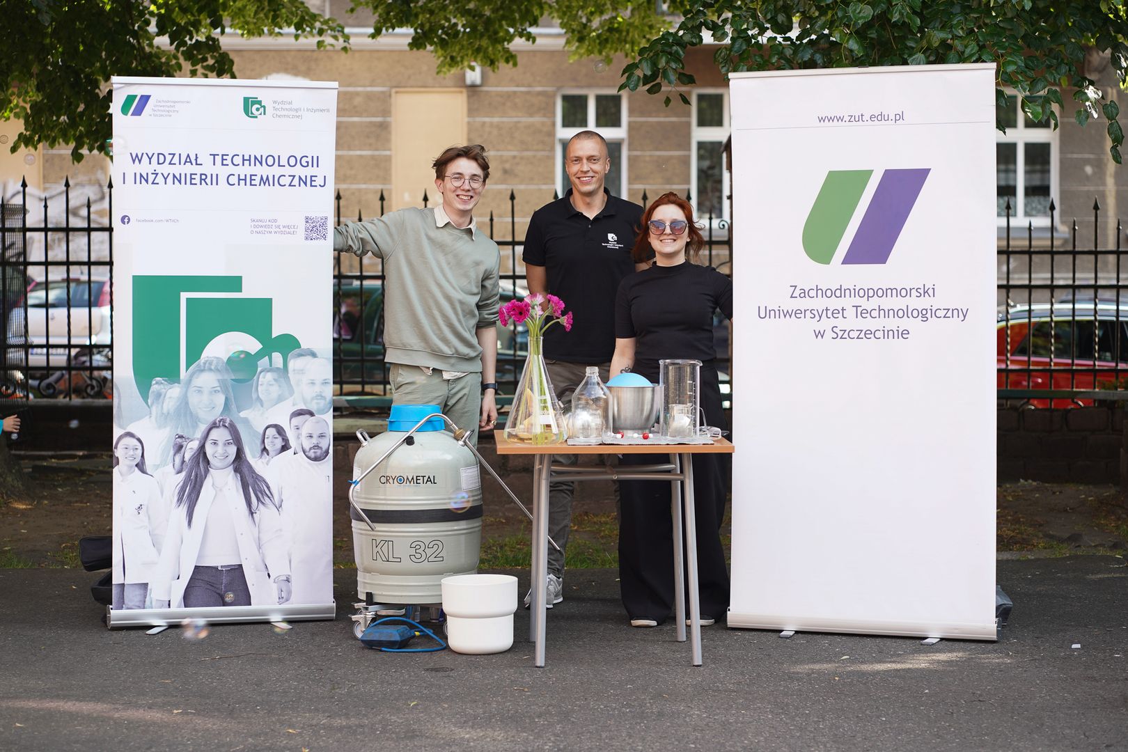 Trzy osoby stoją za stołem z różnorodnym sprzętem naukowym. Po lewej stronie znajduje się baner z napisem ‘Wydział Technologii i Inżynierii Chemicznej’ oraz grafikami związanymi z chemią. Po prawej stronie widoczny jest baner ‘Zachodniopomorski Uniwersytet Technologiczny w Szczecinie’. 