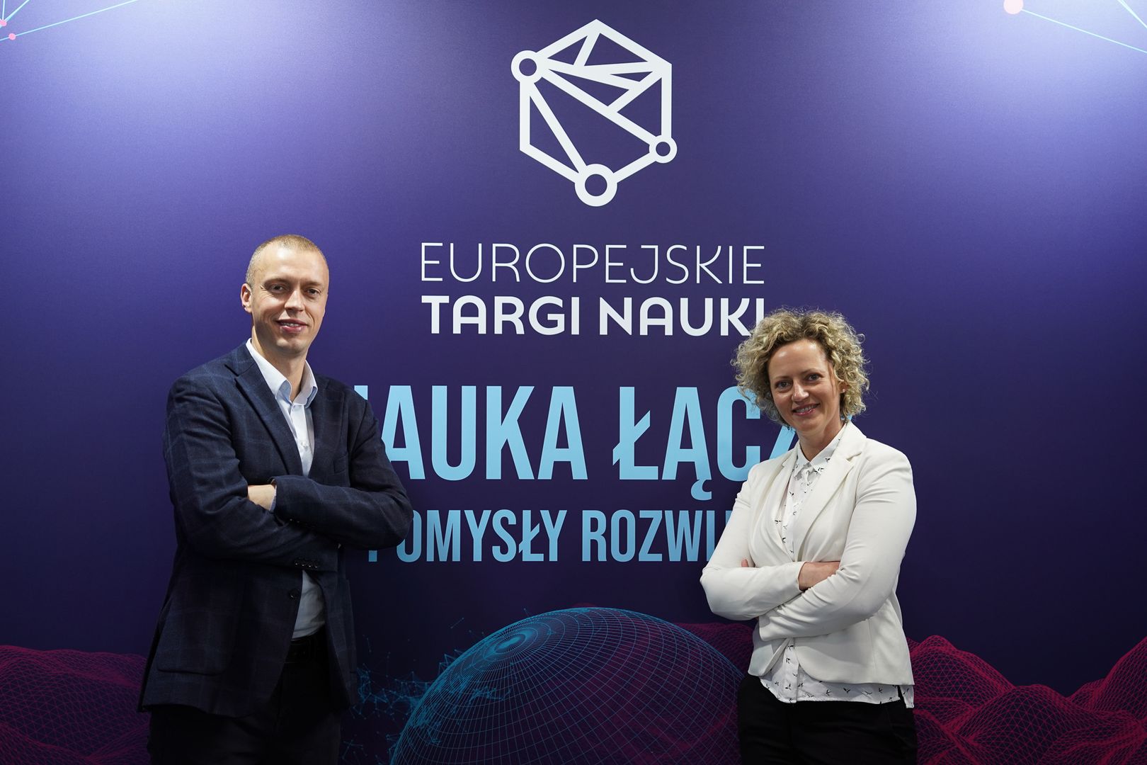 Kobieta i mężczyzna stoją na tle ścianki z napisem 'Europejskie Targi Nauki. Nauka łączy – pomysły rozwijają'. Oboje uśmiechnięci, mają skrzyżowane ręce. Mężczyzna w ciemnym garniturze, kobieta w białej marynarce.
