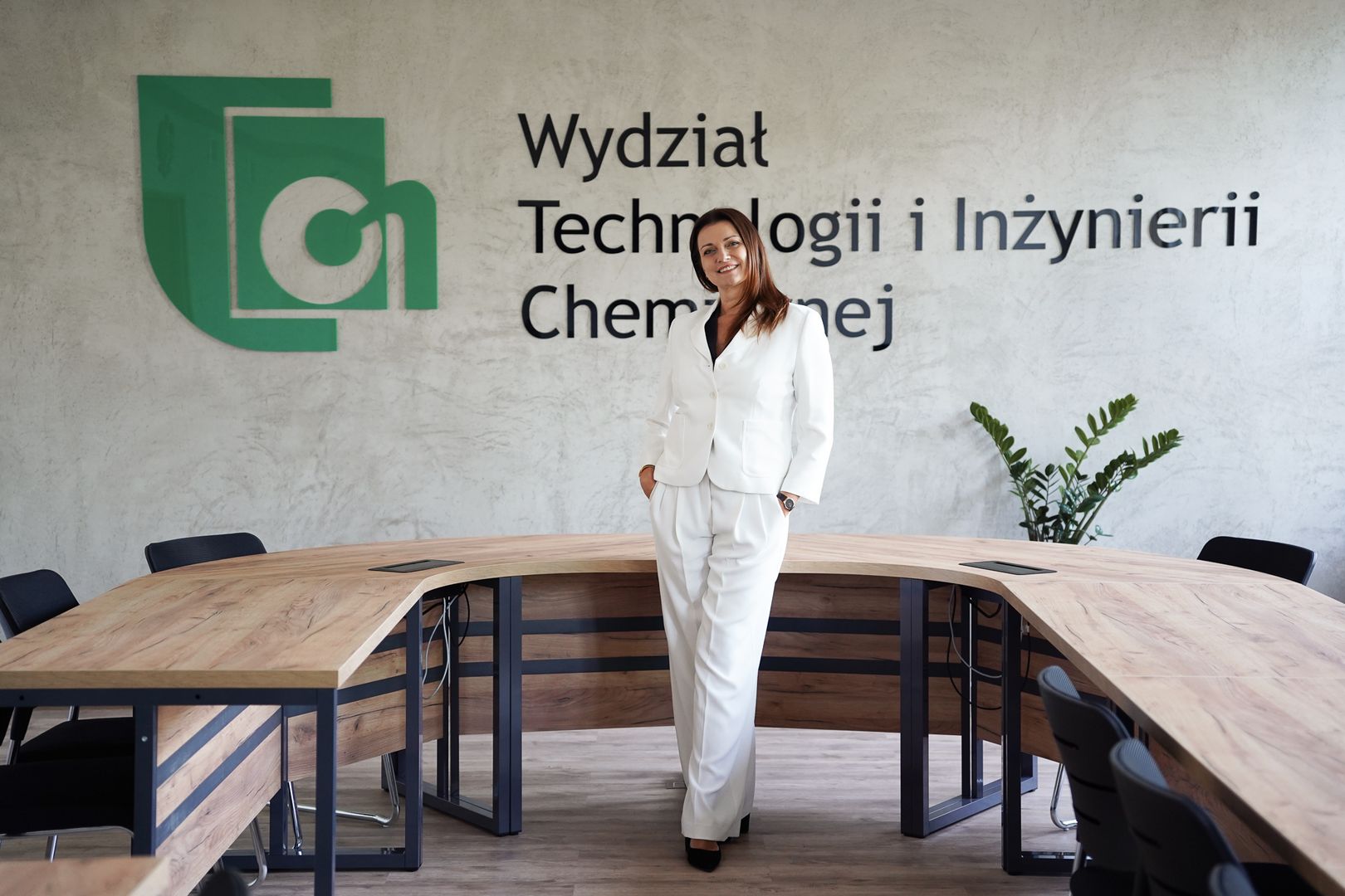 Kobieta w białym garniturze stojąca w sali konferencyjnej Wydziału Technologii i Inżynierii Chemicznej, na tle logotypu i napisu na ścianie.