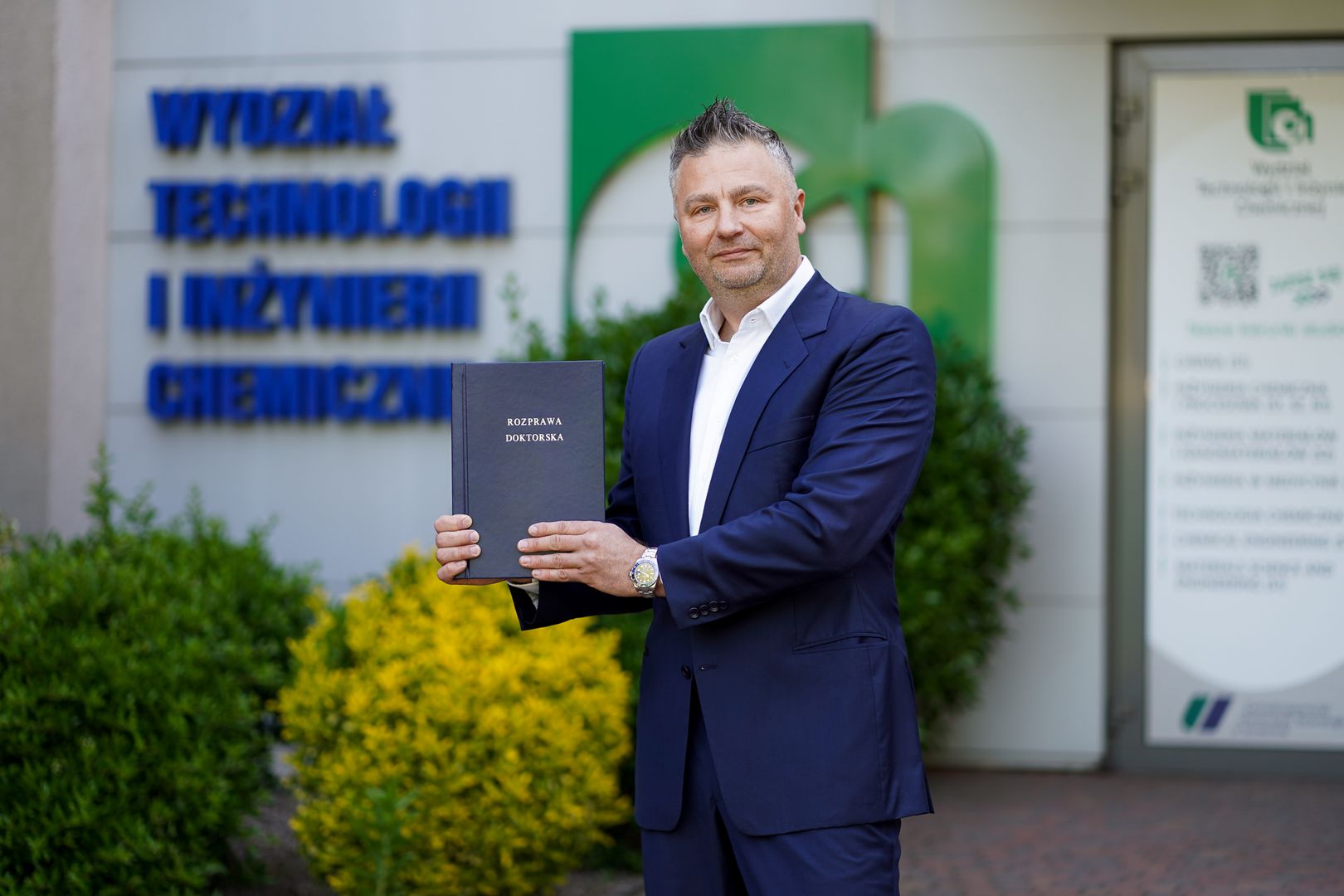 Mężczyzna w garniturze stoi przed budynkiem i trzyma w ręku rozprawę doktorską 
