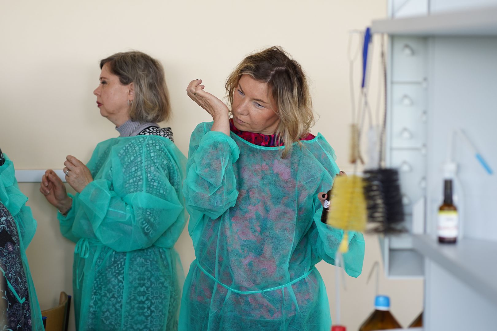 Dwie kobiety w ochronnych fartuchach znajdują się w laboratorium; jedna z nich wącha nadgarstek, podczas gdy druga stoi bokiem. Na pobliskich półkach widoczne jest sprzęt laboratoryjny.
