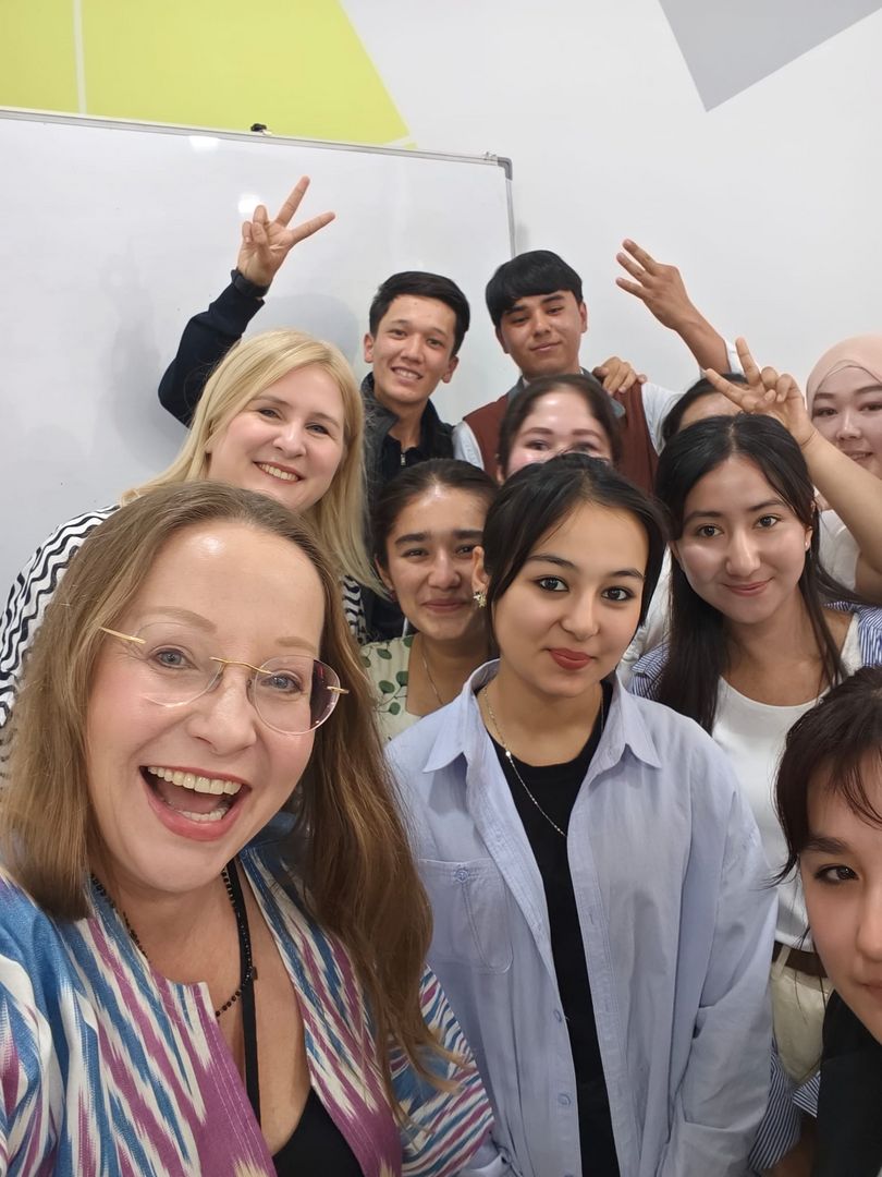 Grupa uśmiechniętych studentów i wykładowczyń podczas spotkania w Karshi Engineering Economic Institute w Uzbekistanie. Na pierwszym planie dwie kobiety, jedna z nich robi selfie. W tle biała tablica i kolorowe ściany.